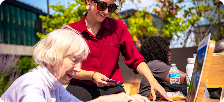 New Arrivals caregiving activity shows a woman assisting an older adult with a colorful tabletop task outdoors.