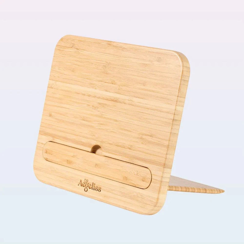 A light natural wood stand with rounded corners and a built-in slot, shown standing upright on a pale blue background. The brand name 'Adelillos' is engraved on the front.